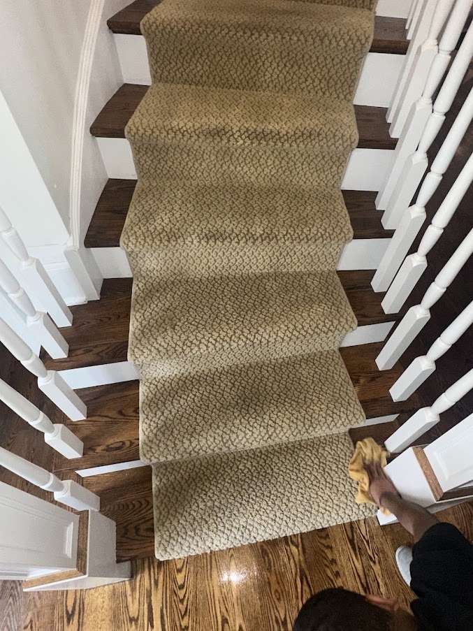 View from above of carpeted stairs with a tan, patterned runner and dark wood edges. A person at the bottom right is cleaning the wooden floor with a yellow cloth, showcasing attention to detail found in professional carpet cleaning. White railings flank both sides.